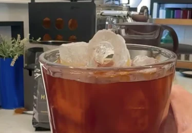 כוס קפה קר — קולד ברו עם קוביות קרח גדולות, על רקע מטחנת קפה בחנות קיקו קפה